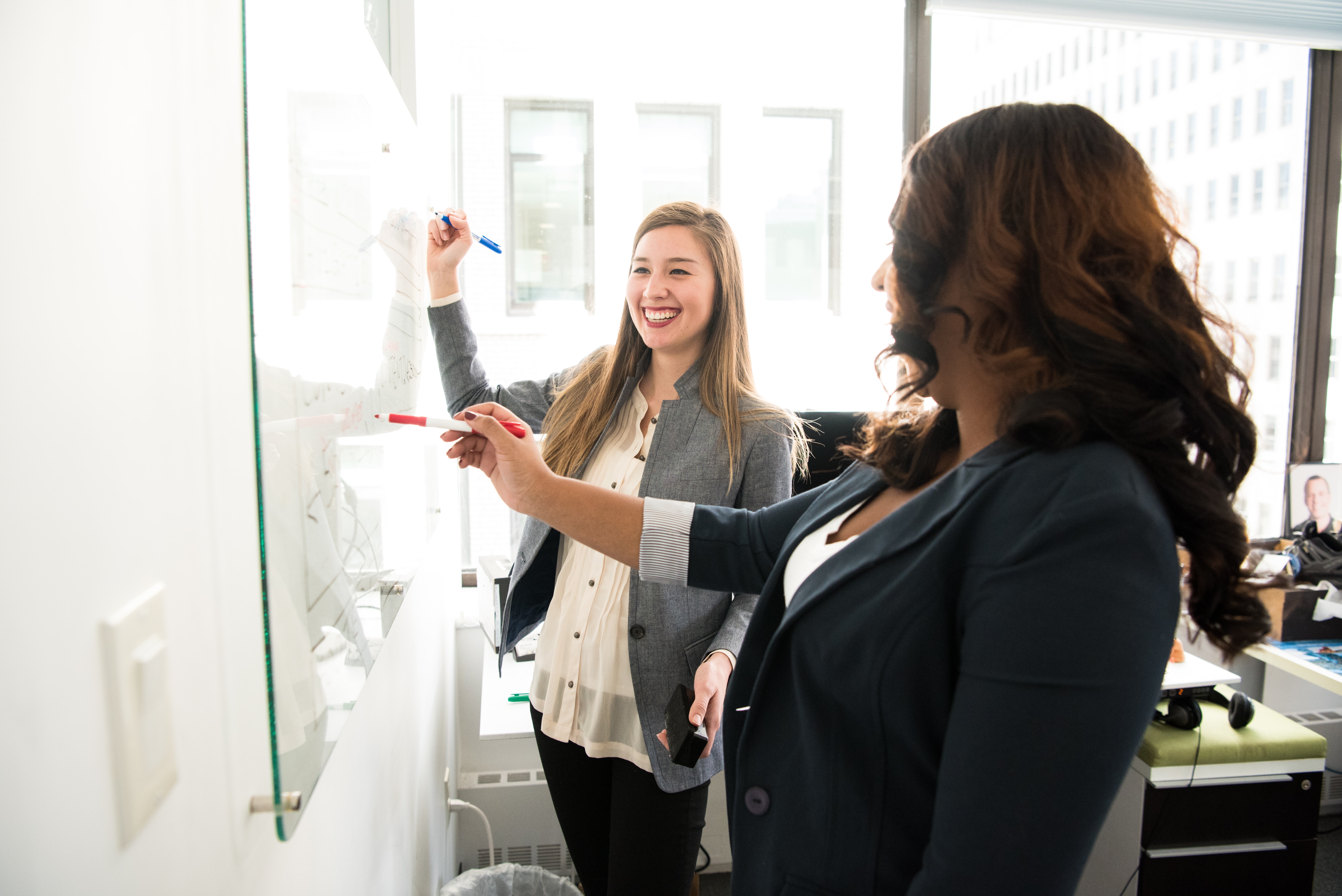 Two smiling woman in office setting writing on white board