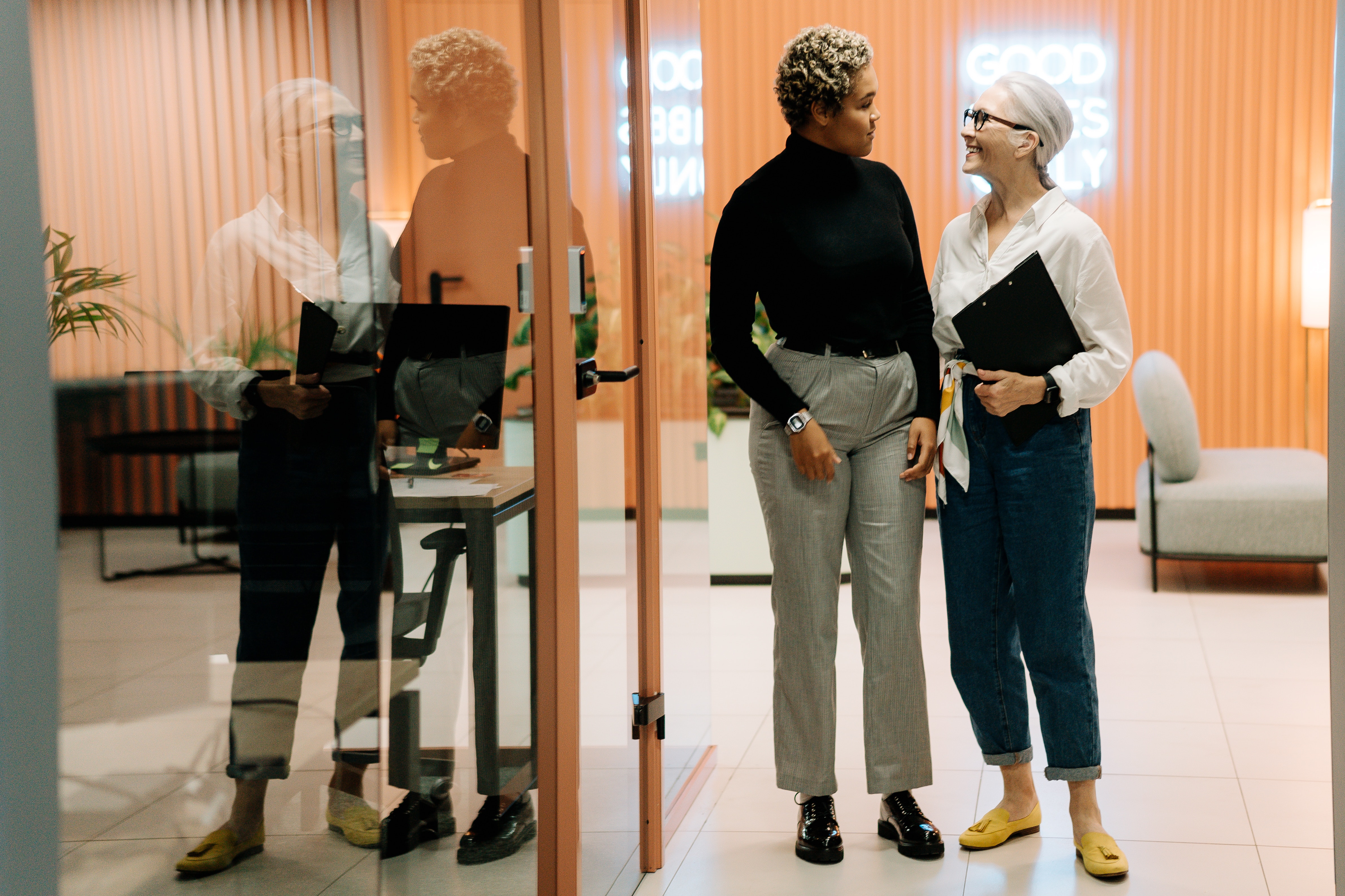 Two females standing in an office corridor smiling at each other