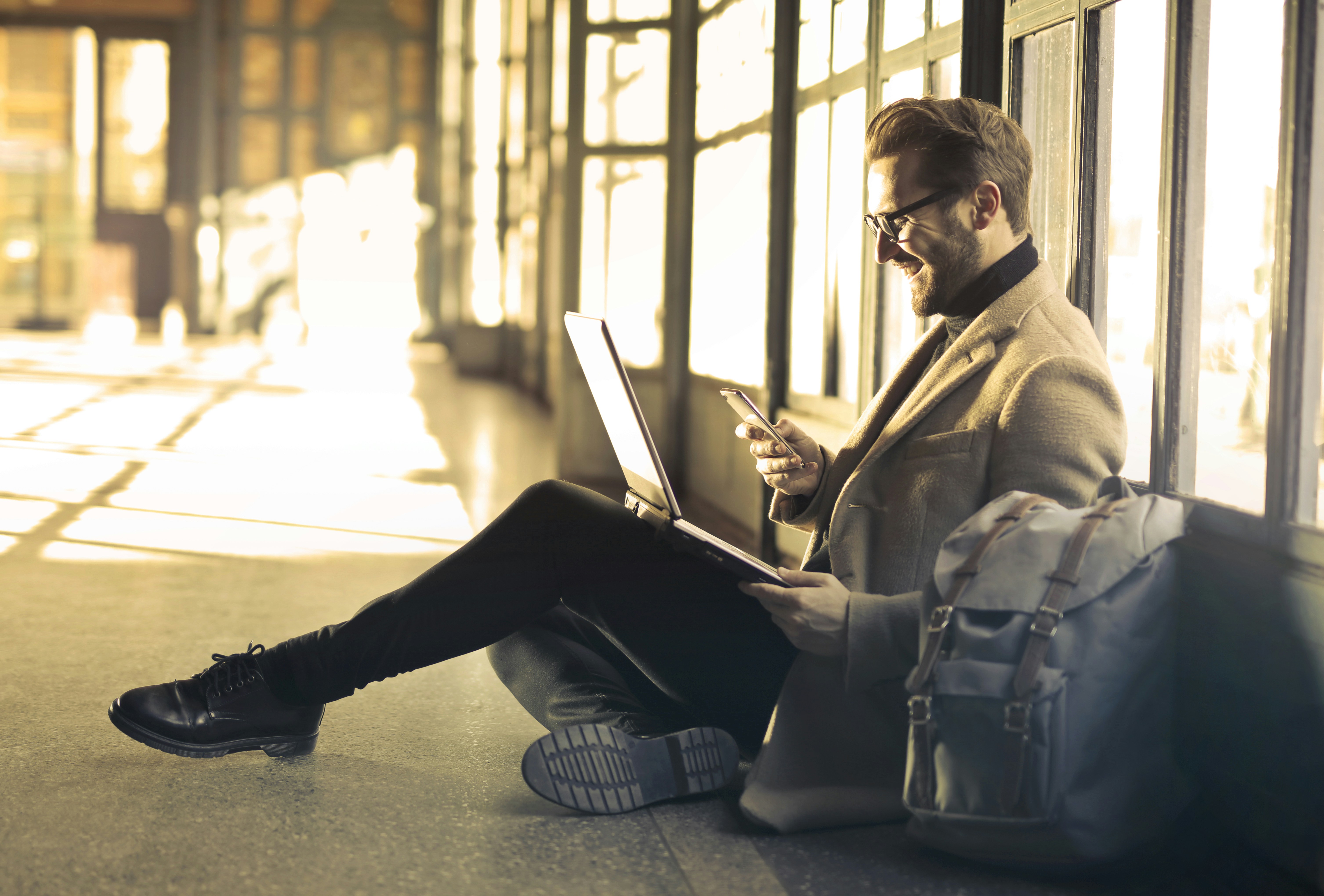 Man sat on floor with laptop on laptop and smiling into mobile phone