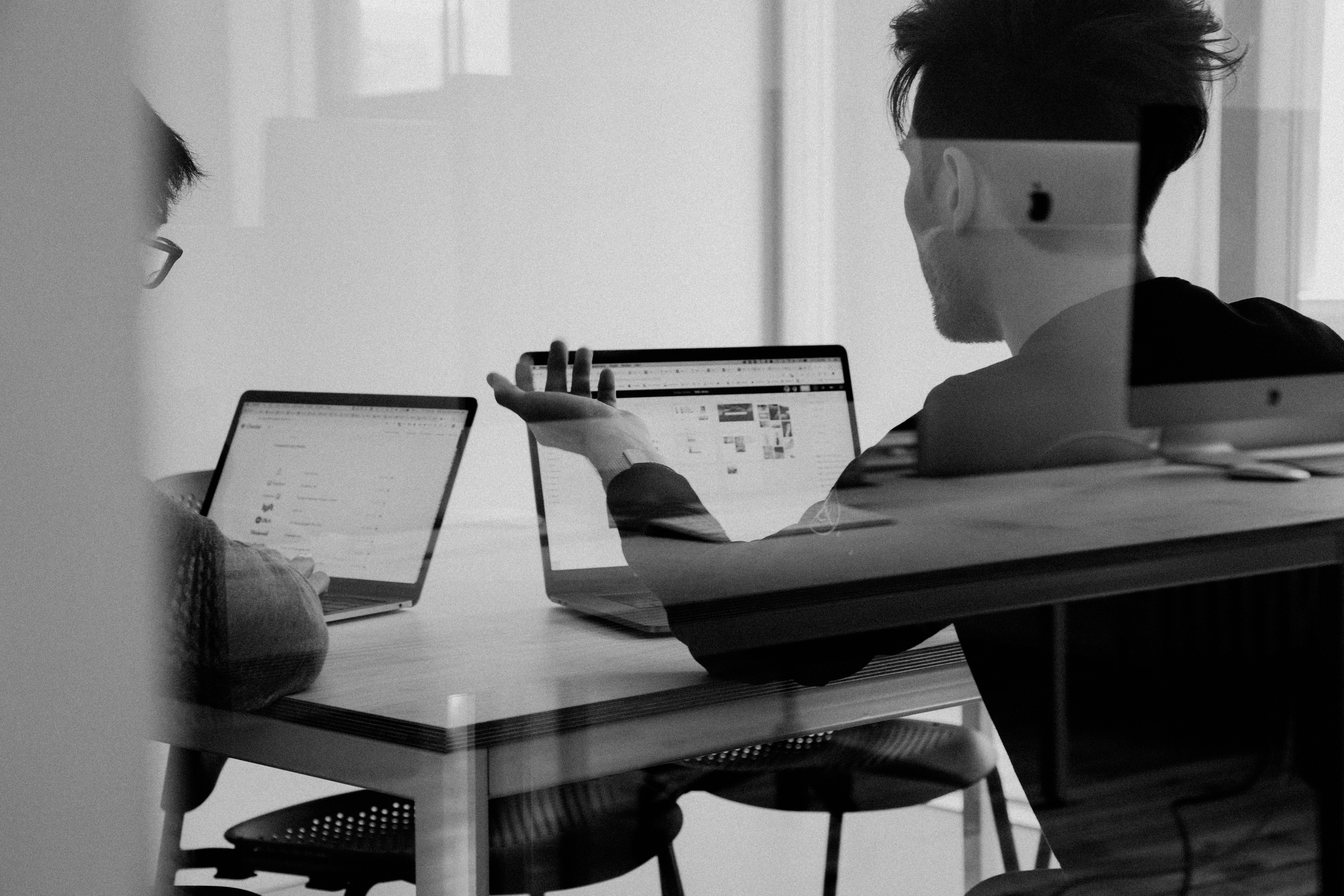 Black and white image taken through window of the back of two people looking at laptop