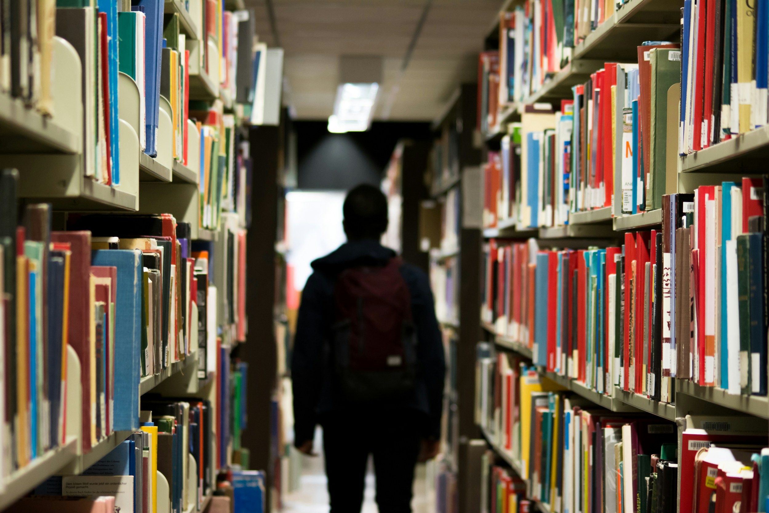A man with a backpack walking through a library with his back to the camera